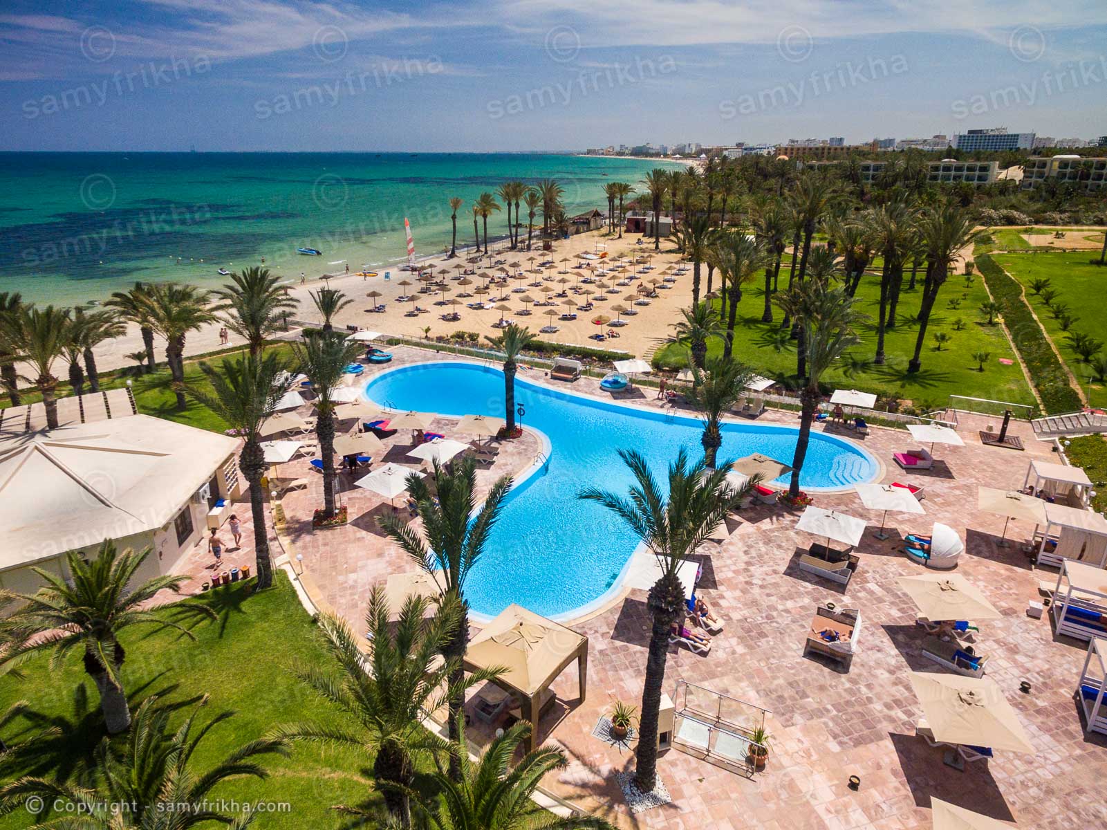 Vue aérienne d'un hôtel avec piscine, jardins et plage en bord de mer.